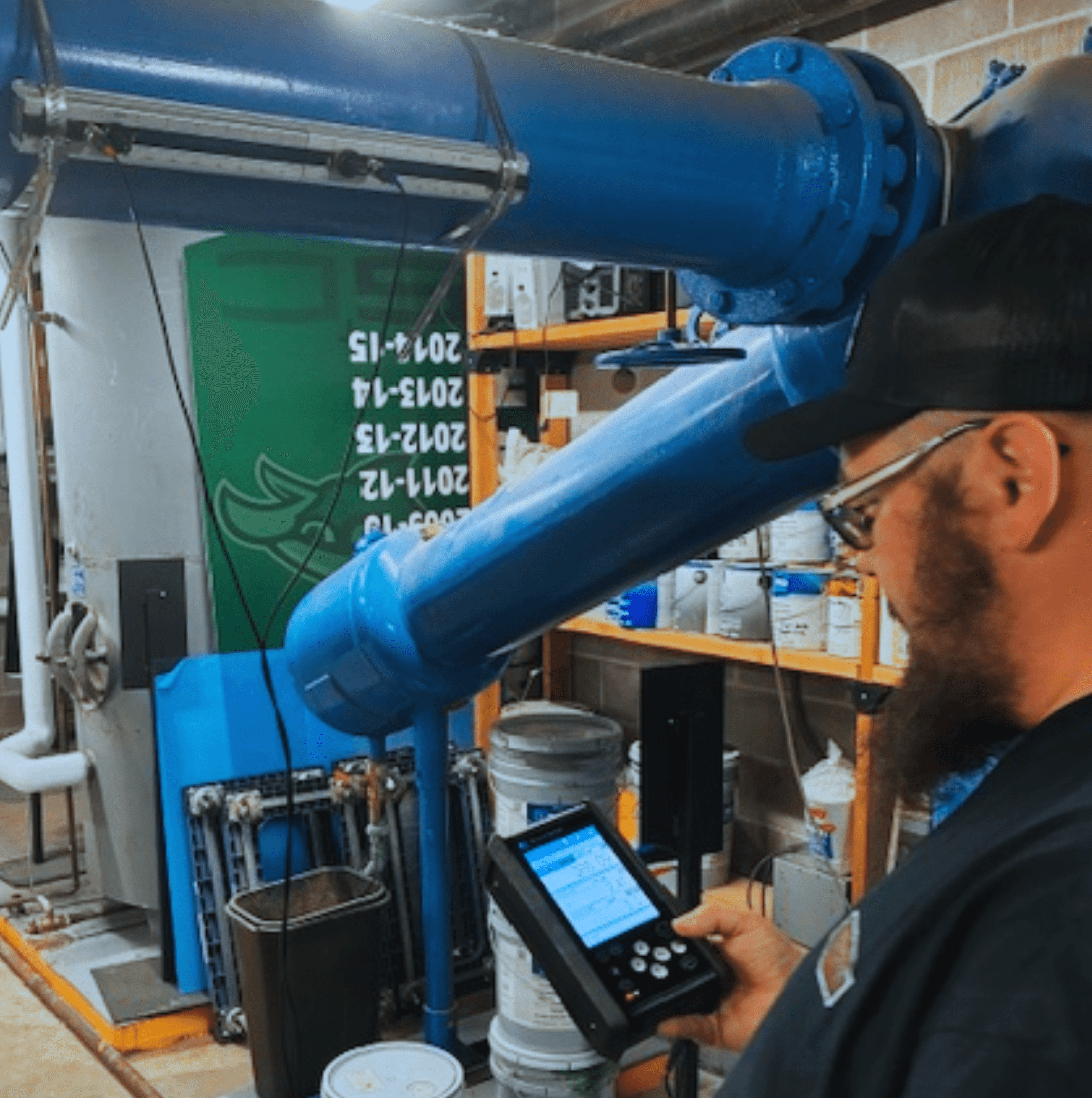 A person operates a control device next to large blue industrial machinery in a workshop filled with equipment and containers.